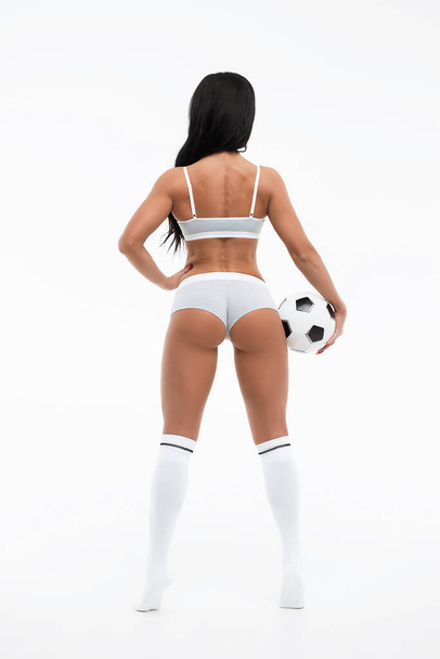 Αθλητική ικανοποιημένη μελαχρινή με λευκά εσώρουχα και κάλτσες στο γόνατο με σφυρίχτρα στο λαιμό και ποδόσφαιρο στο χέρι απομονωμένη σε λευκό φόντο - Φωτογραφία, εικόνα