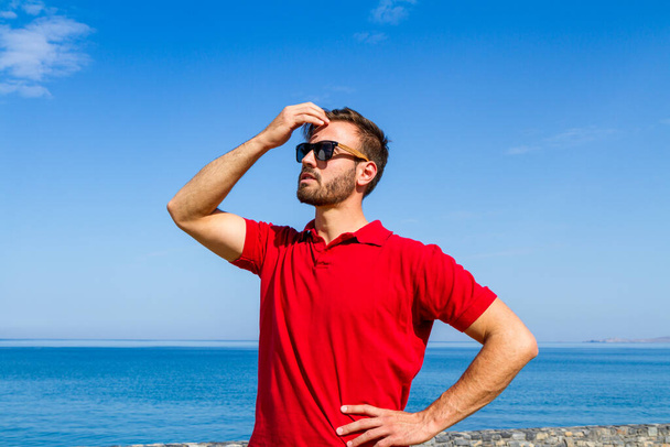 Dinâmico, ambicioso, bem sucedido, expressivo barbudo jovem em vermelho t-shirt óculos de sol no verão azul céu oceano fundo. Conceito de estilo de vida de autoconfiança - Foto, Imagem