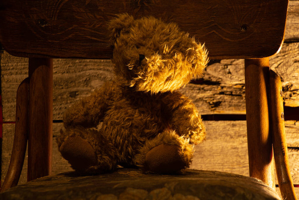 bébé ours en peluche brun assis sur une chaise dans une pièce sombre avec une lampe - Photo, image