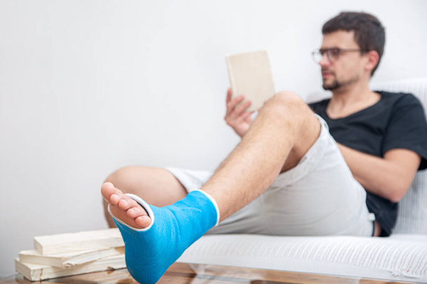Man met gebroken been in blauwe spalk voor de behandeling van verwondingen door enkelverstuiking leesboeken thuis revalidatie. - Foto, afbeelding