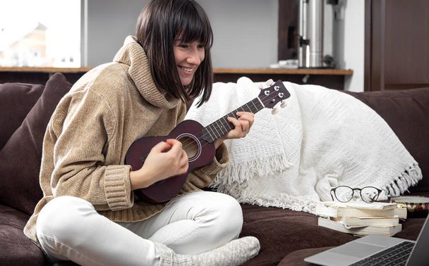 La ragazza allegra giovane in un maglione impara a giocare l'ukulele. Il concetto di apprendimento online, educazione a casa. - Foto, immagini