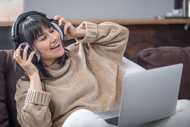 Beautiful girl in headphones listening to music at home on the couch with a laptop. - Фото, изображение