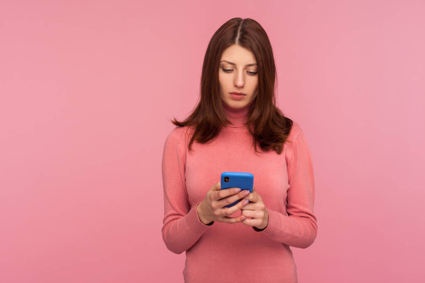 Serious brunette woman holding smartphone in hands, chatting with friends or making food order, online services. Indoor studio shot isolated on pink background - Фото, зображення