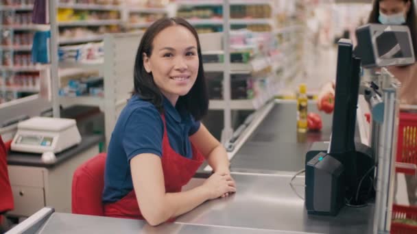 Közepes portré barátságos nő egyenruhában dolgozik pénztárosként nagy szupermarketben mosolyog a kamera előtt, miközben felismerhetetlen ügyfél maszkban termékeket helyez szállítószalagra homályos háttérrel - Felvétel, videó