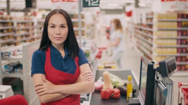 Medium lähikuva muotokuva kaunis sekarodun nainen työskentelee kassanhoitaja hypermarket seisoo kädet taitettu kuljetinhihna täynnä tuotteita taustalla - Materiaali, video