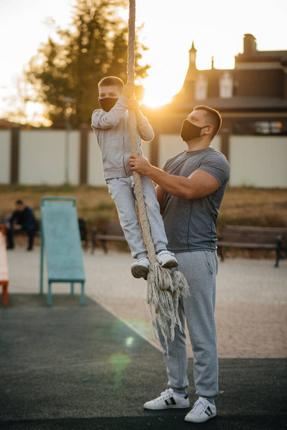 A father helps his son climb a rope on a sports field in masks during sunset. Healthy parenting and healthy lifestyle - Valokuva, kuva