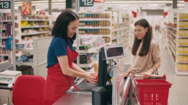 Közepes lövés mosolygós női pénztáros piros egyenruhában beszél vidám nő sorban, miközben mérlegeli zöldségek szállítószalag - Felvétel, videó