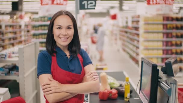Medium close-up portrait of young smiling female cashier wearing red apron standing with hands folded in big mall while people doing shopping in background - Video