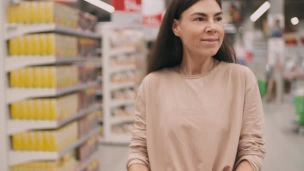 Inclinando-down médio metragem de jovem mulher caucasiana com carrinho fazendo compras diárias sorrindo enquanto procura produtos em grande supermercado - Filmagem, Vídeo