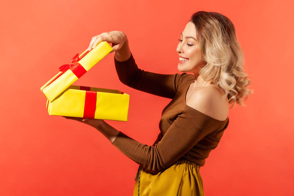 Happy beautiful woman unboxing big yellow present and looking inside with satisfied smile on face, enjoying present. Indoor studio shot isolated on orange background - Foto, immagini