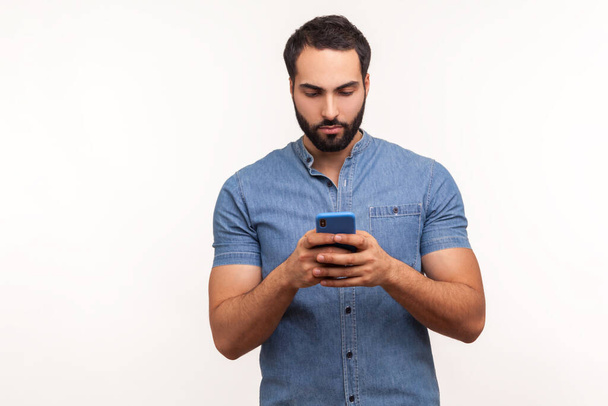 Assertive bearded man in blue shirt attentively looking at display of smartphone he holding, surfing internet, doing shopping online. Indoor studio shot isolated on white background - Foto, immagini
