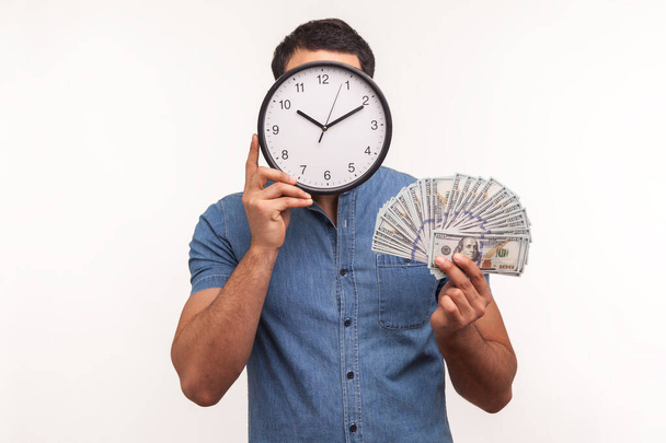 Man in blue shirt hiding behind wall clock holding hundred dollar banknotes in hand, time to make money. Indoor studio shot isolated on white background - Fotó, kép