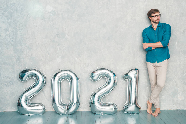 Portrait of smiling handsome man posing near wall. Sexy bearded male staning near Silver 2021 Balloons. Happy New 2021 Year. Metallic numbers 2021 - 写真・画像