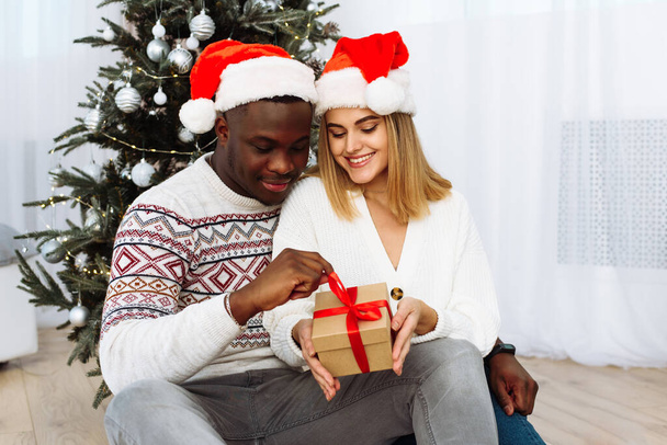 Kochająca się para śmiejąca się, patrząca w kamerę, trzymająca prezent świąteczny i uśmiechająca się w Sylwestra. czarny mężczyzna przytula biały młody kobieta na tle choinki - Zdjęcie, obraz