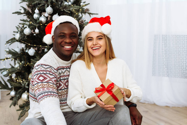Porträt eines lächelnden jungen Paares, das neben dem Weihnachtsbaum sitzt und gemeinsam das neue Jahr feiert. Glückliche Männer und Frauen genießen Winterfest an geschmücktem Tannenbaum - Foto, Bild