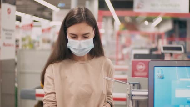 Medium close-up of young woman wearing disposable mask standing in queue waiting for her products being scanned by cashier in hypermarket - Metraje, vídeo