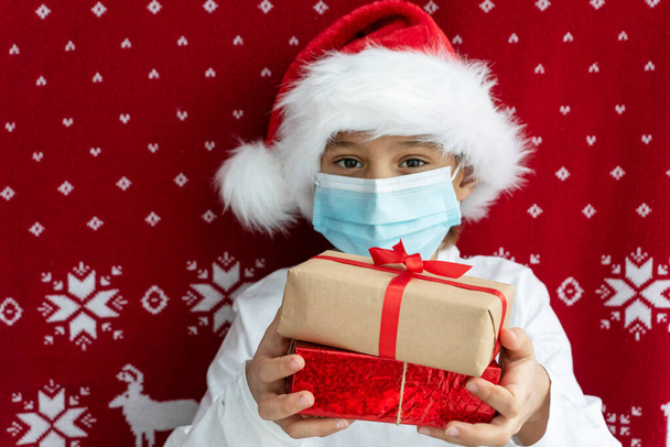 ragazzo in cappello di Babbo Natale e maschera medica viso tiene due scatole regalo stanco con nastri su sfondo rosso di Natale. Concetto vacanza - Foto, immagini