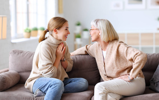 Die erwachsene Tochter gestikuliert und spricht mit einer fröhlich lachenden älteren Mutter, während sie sich zu Hause auf der Couch ausruht. - Foto, Bild