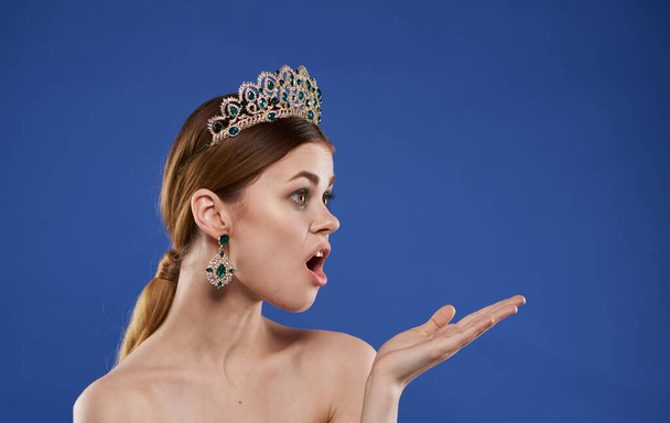 A woman with bare shoulders and a diadem on her head gestures with her hands on a blue background - Fotoğraf, Görsel