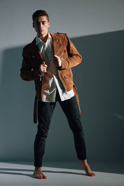 Attractive young man in a brown jacket and trousers bent over to the side indoors against a gray background - 写真・画像
