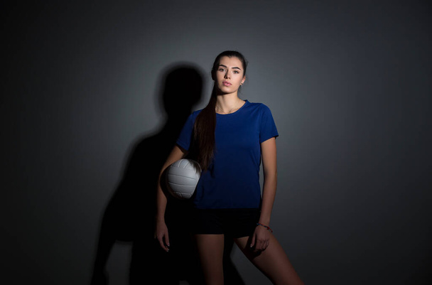 Volleyball girl hold and kick ball in blue costume on dark wall background. Player doing sport workout at home. Sport and recreation concept - Фото, изображение