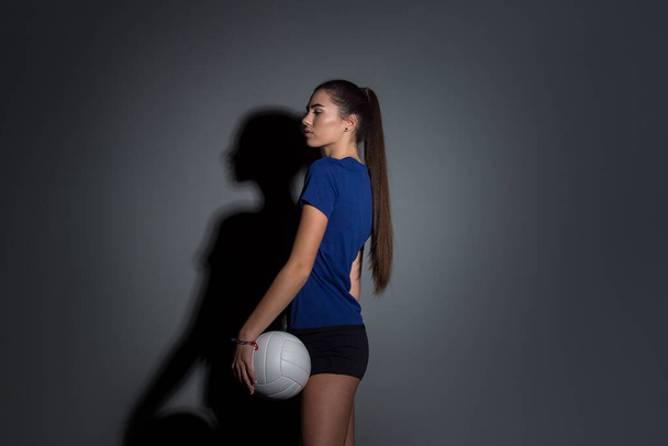 Volley-ball fille tenir et coup de pied boule en costume bleu sur fond de mur sombre. Joueur faisant de l'entraînement sportif à la maison. Concept de sport et de loisirs - Photo, image