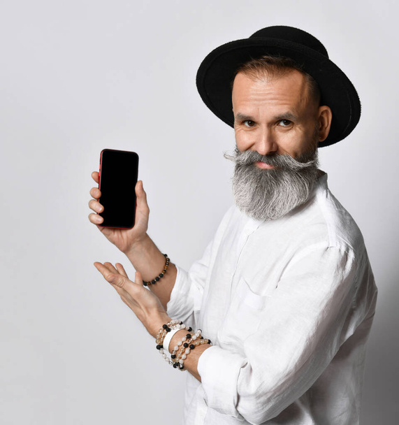Stylish hipster man in black hat looking at camera pointing at cellphone he is holding in hand. - Foto, imagen