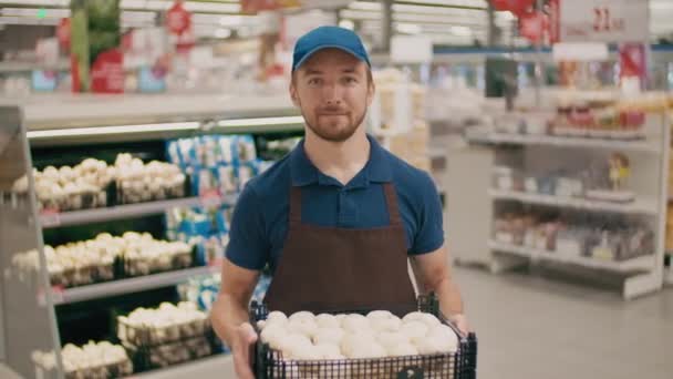 Portrait moyen d'un beau vendeur d'épicerie en uniforme souriant à la caméra tenant un grand panier de champignons blancs - Séquence, vidéo