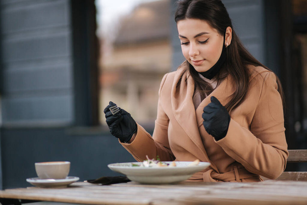 Hermosa joven almorzando en la cafetería al aire libre en la terraza. Mujer con máscara protectora negra. Mujer quitarse la máscara al comer - Foto, Imagen