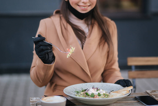 Großaufnahme einer Frau in schwarzen Handschuhen, die Besteck hält. Cafe-Konzept während der Qurantine. Zerrissen auf der Gabel. Essen im Freien in Schutzhandschuhen. Frischer veganer Salat mit Garnelen - Foto, Bild