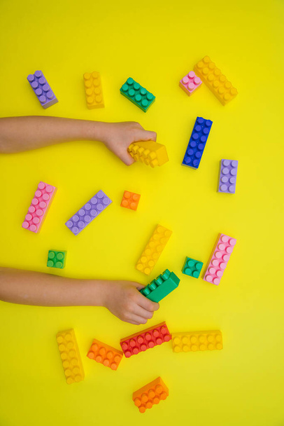Children's hands hold the details of the constructor on a yellow background. Multi-colored construction blocks. Children's educational game. Background and texture. - 写真・画像