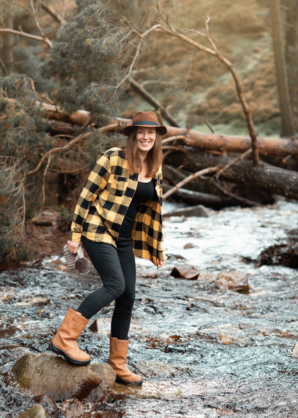 mujer en camisa amarilla y sombrero caminando a lo largo del río en el bosque de otoño - Foto, imagen