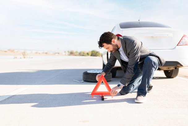 Latinský muž umísťující reflexní trojúhelník na silnici jako varování, protože jeho auto má píchlou pneumatiku  - Fotografie, Obrázek