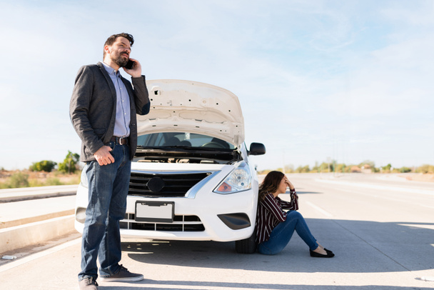 Hombre hispano hablando por teléfono con un mecánico. Mujer joven mirando molesto y sentado junto a un coche con la capucha abierta - Foto, imagen