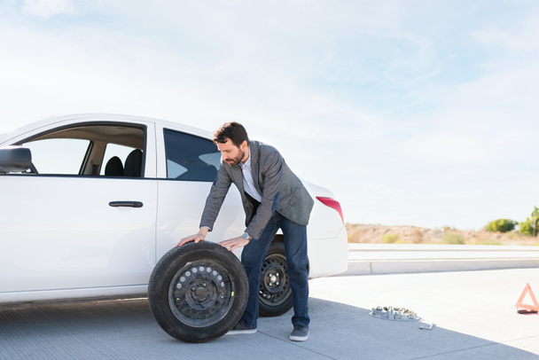 Latin man pulling the spare tire out of his car to change the flat one on the roadside - Foto, immagini