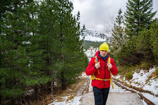 Kırmızı ceketli, sarı şapkalı ve sırt çantalı yakışıklı bir turist dağ yolunda duruyor.. - Fotoğraf, Görsel