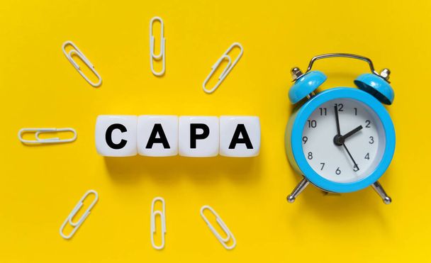 Finance and economics concept. On a yellow background, a blue alarm clock, paper clips and white cubes on which the text is written - CAPA - Фото, изображение