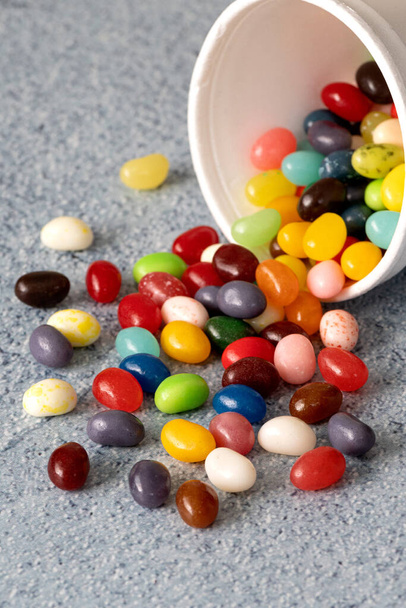  Jelly beans que se derrama de la taza de espuma blanca contra la mesa de fondo azul, composición festiva y colorida con candie, vista lateral con una sensación dinámica  - Foto, Imagen