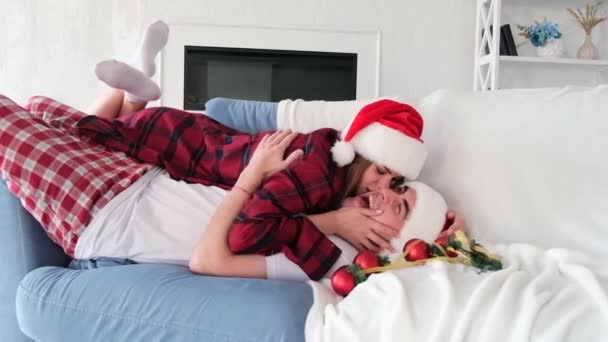 Mujer joven juguetona se encuentra en la parte superior del hombre besándolo y tonteando en el sofá rodeado de decoraciones de árboles de Navidad. Pareja vistiendo sombreros de santa y pijamas a cuadros rojos - Metraje, vídeo