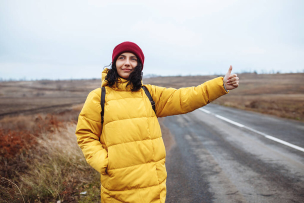 Una turista con una mochila con chaqueta amarilla y sombrero rojo coge un coche en la carretera. Mujer joven viaja durante la temporada de invierno o finales de otoño. Hitch-senderismo, viaje, pulgar hacia arriba, concepto de viaje - Foto, imagen
