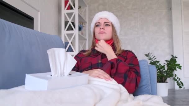 Colds, quarantine and healthcare concept. Woman puts protective medical mask on her face lying at home on sofa under blanket in red pajamas and santas hat, woman has acold and is not feeling well - Footage, Video
