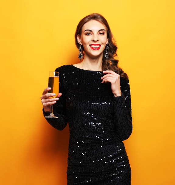 Young stylish woman drinking champagne, celebrating new year, wearing evening dress. Happy New Year to you. One young and beautiful woman dancing with glass of champagne and smiling. - Фото, зображення