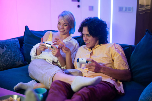 Gelukkige jongeren spelen videospelletjes op de console terwijl ze op de bank voor de tv zitten. Millennial koppel besteden leuke tijd samen thuis. Kamer met neonverlichting. - Foto, afbeelding