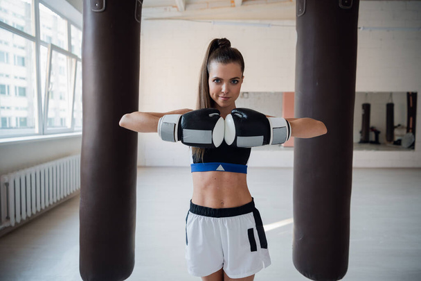 Симпатичная девушка позирует для фото в боксёрских перчатках на фоне боксёрских груш - Фото, изображение