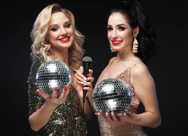 Two women in evening dresses, chic hairstyles and makeup, sing with a microphone, holding a disco ball - Φωτογραφία, εικόνα