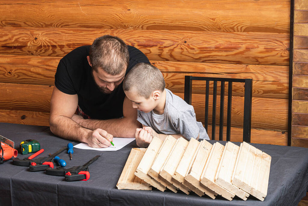 Pai e filho estão desenhando em papel, planejando como construir uma casa de pássaros, ferramentas e uma viga na mesa na oficina. Conceito de treinamento de carpintaria para crianças - Foto, Imagem