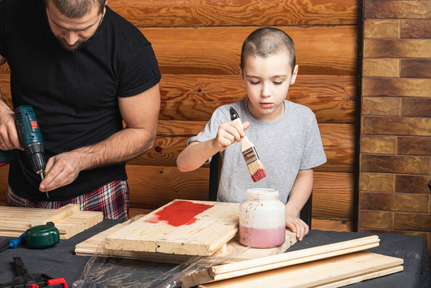 Papà e figlio stanno dipingendo una tavola di legno con un pennello in rosso, come costruire una casa di uccelli, strumenti e una trave sul tavolo in officina. Concetto di formazione falegnameria per bambini - Foto, immagini