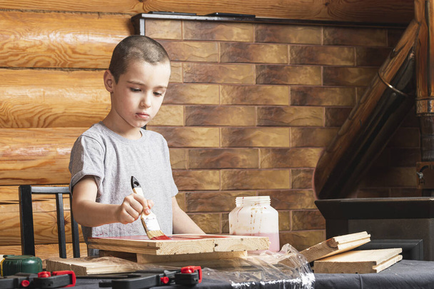 Um menino pinta uma falsificação de madeira com um pincel em vermelho em uma casa de campo. Pintar as paredes. Rapaz a fazer reformas em casa. - Foto, Imagem