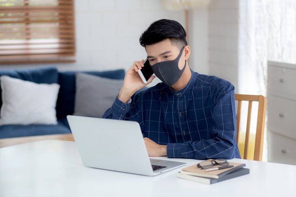 顔のマスクの若いアジアのビジネスマンは、スマートフォンを話し、ノートパソコン上で動作するcovid-19を保護するために、ビジネスマンの隔離作業ノートブック、社会的距離と自宅から家に滞在. - 写真・画像