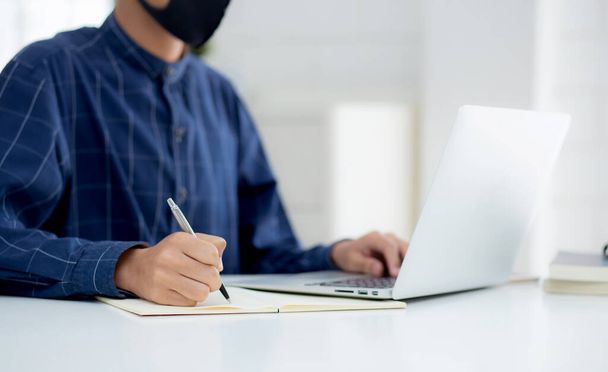 顔のマスクで若いアジアのビジネスマンは、 covid-19を保護し、ノートパソコン上で動作し、ノートブックを書く、家からビジネスマン隔離作業、新しい通常、社会的距離と家にとどまる. - 写真・画像